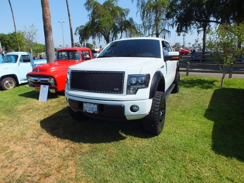 2003 Ford Crown Victoria - white customized Ford F-150 with a blacked-out mesh grille parked next to a red 1950s Ford F-100 at a car show.