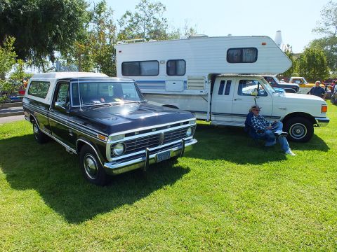 2003 Ford Crown Victoria - black 1970s Ford pickup with a white canopy parked beside a fifth-wheel camper trailer, a man seated nearby on the grass.