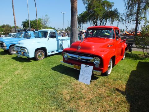 2003 Ford Crown Victoria - bright red 1950s Ford F-100 stepside displayed beside a light blue Ford pickup on show grass with a printed info card.