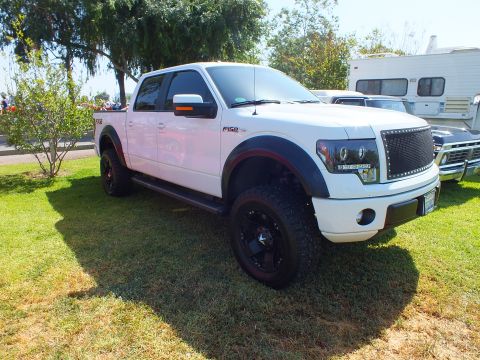 2003 Ford Crown Victoria - white lifted Ford F-150 FX4 crew cab on black off-road wheels parked on show grass with RVs in the background.