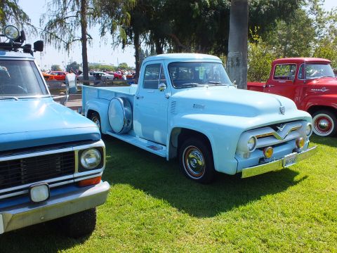 2003 Ford Crown Victoria - light blue 1955 Ford F-100 stepside with a side-mounted spare parked next to a 1970s Ford pickup and a red truck at a show.