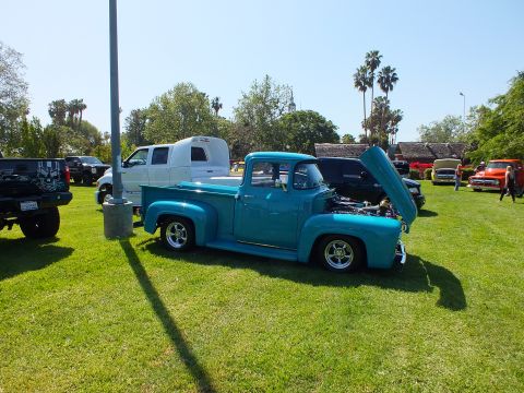 2003 Ford Crown Victoria - teal-and-blue 1956 Ford F-100 stepside with its hood raised, parked on show grass with palms and other vehicles behind.