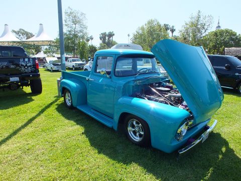 2003 Ford Crown Victoria - glossy teal 1956 Ford F-100 stepside displayed hood-up on show grass, revealing a detailed polished engine bay.