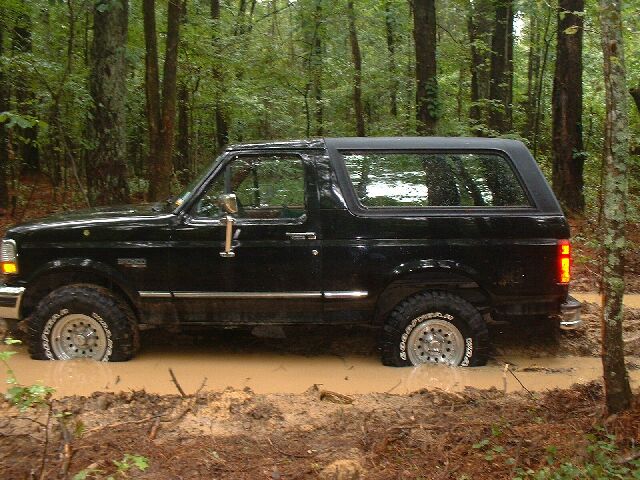 Black Ford Bronco stuck in mud, side view. The vehicle is partially submerged in a muddy trail surrounded by trees