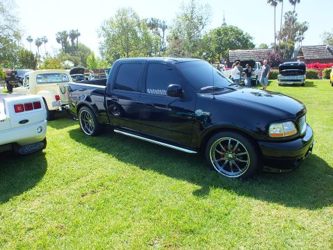 2003 Ford Crown Victoria - glossy black crew-cab pickup with custom chrome wheels is displayed on a lawn alongside other vehicles and spectators.