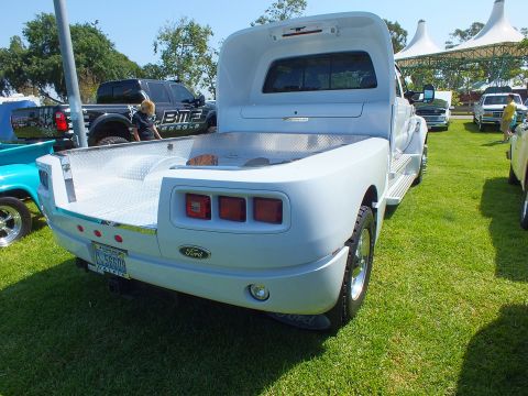 2003 Ford Crown Victoria - white custom dually with a diamond-plate flatbed and Ford tailgate badge is parked rear-facing on a grassy show field.