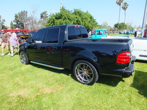 2003 Ford Crown Victoria - black lowered crew-cab pickup with polished chrome wheels and red tail lights sits on a show lawn among other vehicles.