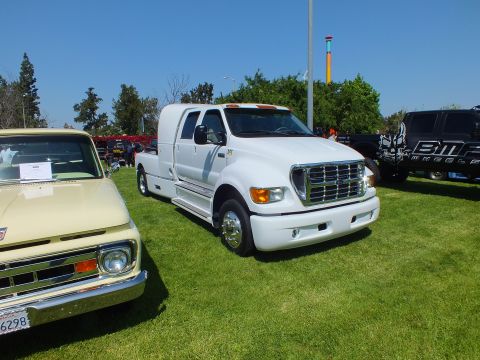 2003 Ford Crown Victoria - white custom crew-cab dually hauler with a chrome grille and running boards is displayed on grass beside a cream vintage pickup.