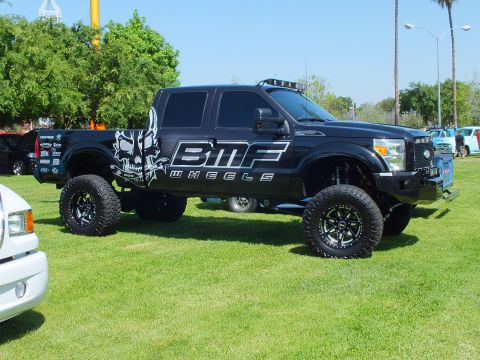2003 Ford Crown Victoria - black lifted crew-cab truck wrapped with BMF Wheels skull graphics, off-road tires, and a roof light bar is parked on grass.