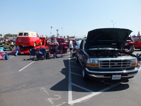2003 Ford Crown Victoria - dark Ford SUV with its hood raised sits in a paved parking-lot car show, with a red vintage panel truck and crowds nearby.