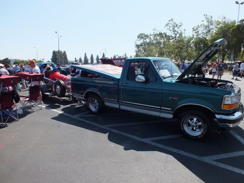 2003 Ford Crown Victoria - green Ford F-150 pickup with its hood up is parked at a paved car show beside a red open-wheel hot rod and crowds of people.