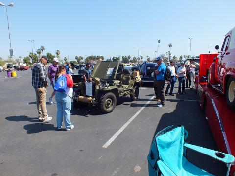2003 Ford Crown Victoria - olive-drab military-style jeep draws onlookers at a paved car show, with a red truck and palm trees in the background.