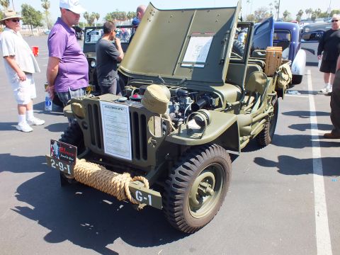 2003 Ford Crown Victoria - olive-drab military jeep with raised hood, exposed engine, and a coiled rope across the front grille sits at a paved car show.