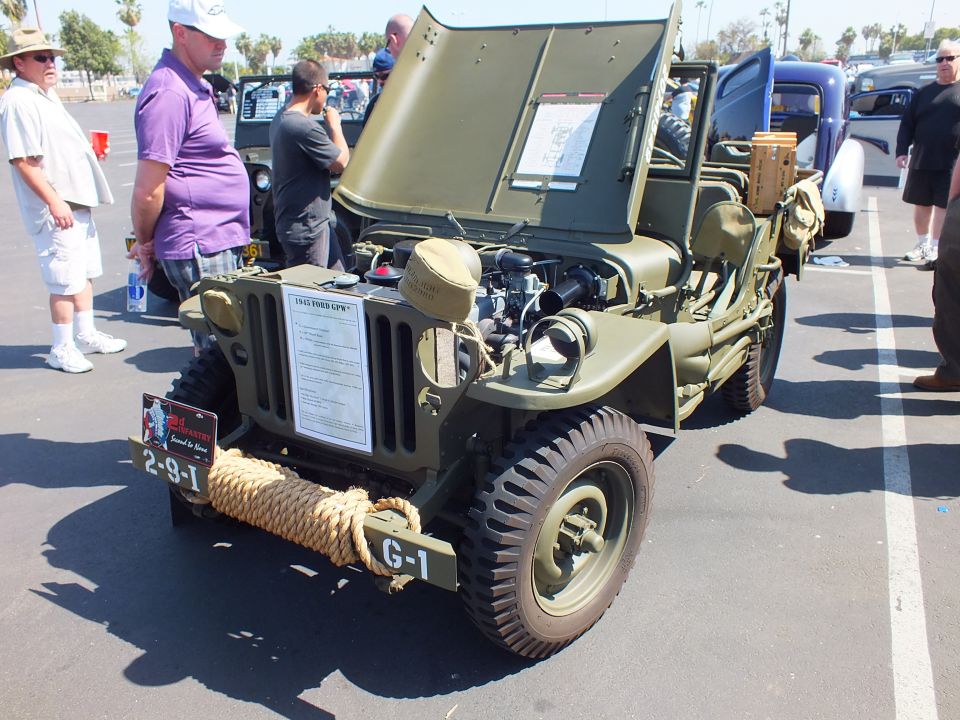 Olive-green military jeep with open hood and rope on front at car show