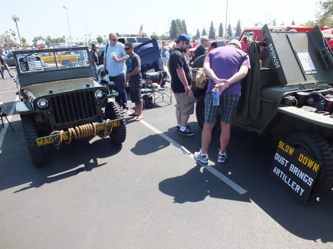 2003 Ford Crown Victoria - Two olive-drab military jeeps are displayed at a paved car show, one with a spare-tire sign reading SLOW DOWN DUST BRINGS ARTILLERY.