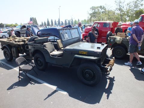 2003 Ford Crown Victoria - olive-drab military jeep marked USMC sits hitched to a gear-laden cargo trailer at a paved car show among other vehicles.