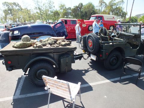 2003 Ford Crown Victoria - olive-drab military jeep with a spare tire tows a trailer loaded with packs and gear at a paved car show under leafy trees.