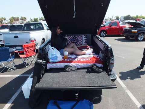 2003 Ford Crown Victoria - woman reclines on patriotic bedding laid in the open bed of a black pickup at an outdoor car show.