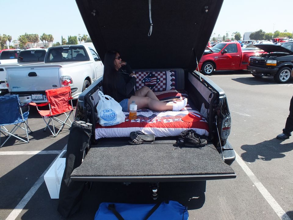 Woman lounging on bedding in the open tailgate bed of a black pickup at a car show