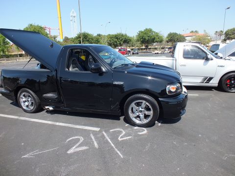 2003 Ford Crown Victoria - black single-cab sport pickup with hood and door open sits at a car show, chalk number 21123 marked on the asphalt.