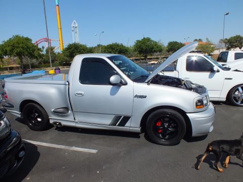 2003 Ford Crown Victoria - silver single-cab sport pickup with hood raised and black wheels at an outdoor car show, with a small dog walking past.