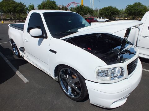 2003 Ford Crown Victoria - white single-cab sport pickup with large chrome wheels and the hood raised, parked at an outdoor car show.