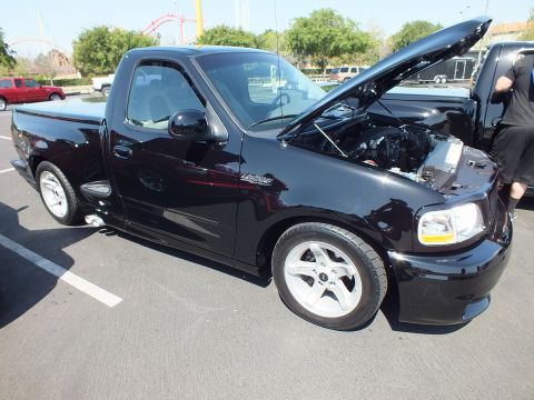 2003 Ford Crown Victoria - lowered black Ford SVT Lightning single-cab pickup with its hood raised, parked on asphalt near a roller coaster.