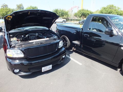 2003 Ford Crown Victoria - Two black Ford pickups parked together at a car show, the nearer one with its hood up and a California plate.