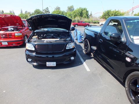 2003 Ford Crown Victoria - row of black and red Ford pickups at an outdoor show, the front black truck with hood up and billet grille.