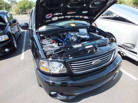 2003 Ford Crown Victoria - black Ford pickup with its hood raised, revealing a supercharged engine and many sponsor stickers underneath.