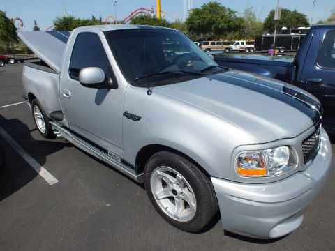 2003 Ford Crown Victoria - silver Ford SVT Lightning single-cab pickup parked on asphalt at an outdoor show beside a roller coaster.