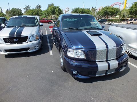 2003 Ford Crown Victoria - dark blue pickup with silver racing stripes parked beside a silver striped Ford pickup at an outdoor show.