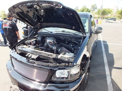 2003 Ford Crown Victoria - black Ford F-150 pickup with hood raised, displaying a chrome billet grille and supercharged engine bay at a show.
