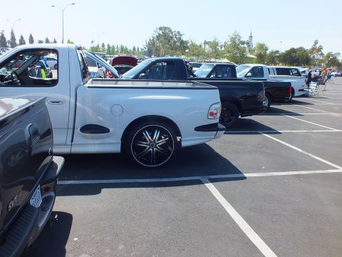 2003 Ford Crown Victoria - white pickup with black-and-chrome wheels leads a row of trucks parked across a sunny parking-lot car show.