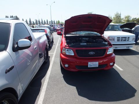 2003 Ford Crown Victoria - red Ford F-150 pickup with its hood raised showing the engine bay, parked beside a silver pickup at an outdoor show.