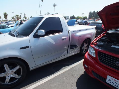 2003 Ford Crown Victoria - white Ford F-150 Lightning regular-cab pickup parked next to a red Ford pickup with its hood open at a busy car show.