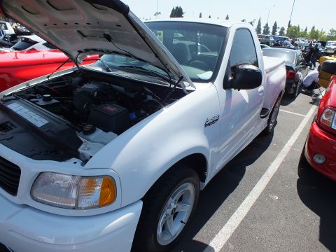 2003 Ford Crown Victoria - Side three-quarter view of a white Ford F-150 Lightning pickup with raised hood at an outdoor car show.
