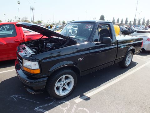 2003 Ford Crown Victoria - black Ford F-150 Lightning pickup with its hood raised parks among colorful trucks at a sunny outdoor car show.
