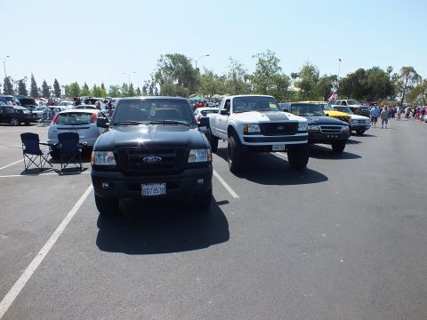 2003 Ford Crown Victoria - black Ford Ranger and a white lifted Ford pickup sit front-to-camera in a row of trucks at a daytime car show.