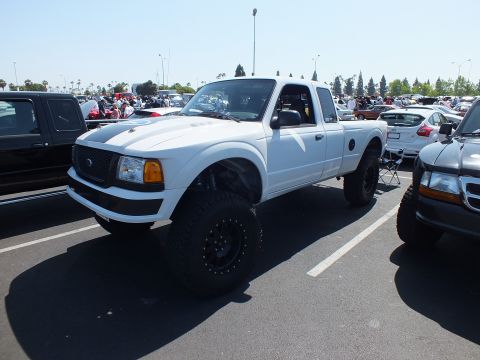 2003 Ford Crown Victoria - white lifted Ford Ranger extended-cab pickup with off-road tires sits among rows of vehicles at a sunny car meet.