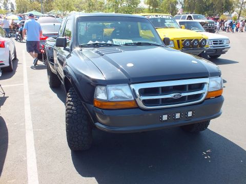 2003 Ford Crown Victoria - dark gray Ford Ranger pickup with chunky tires and a front light bar faces the camera at a crowded outdoor car show.