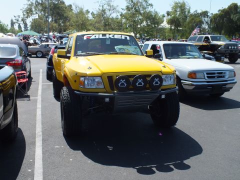 2003 Ford Crown Victoria - yellow Ford Ranger prerunner with a Falken windshield banner and round off-road lights sits in a busy car-show lot.
