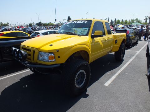 2003 Ford Crown Victoria - yellow lifted Ford Ranger prerunner with front off-road lights and a Falken banner sits among rows of show cars.