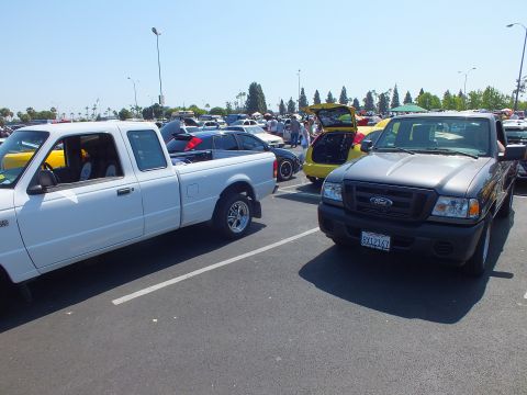 2003 Ford Crown Victoria - white Ford Ranger and a gray Ford Ranger park side by side amid a crowded daytime car-show parking lot.