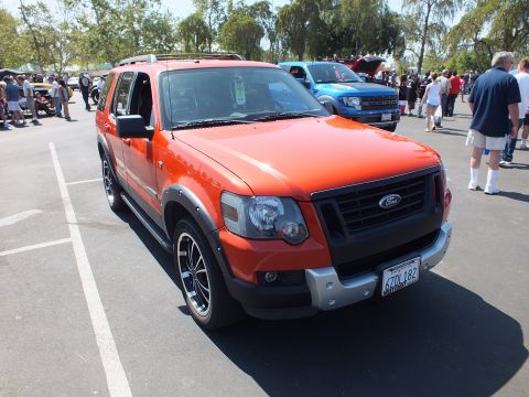 2003 Ford Crown Victoria - two-tone red and black Ford Explorer SUV with a blacked-out grille parks among crowds at a sunny outdoor car show.