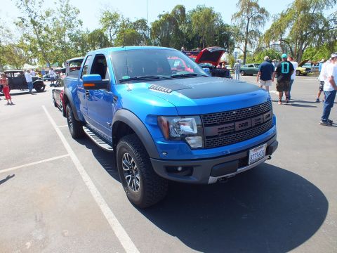 2003 Ford Crown Victoria - blue Ford F-150 SVT Raptor extended cab on knobby off-road tires sits among show vehicles in a sunny parking lot.