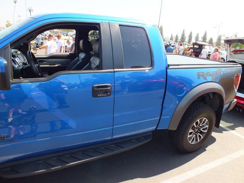 2003 Ford Crown Victoria - blue Ford Raptor pickup is shown from the side with its door open and a Raptor decal along the bedside at a car show.
