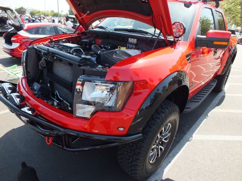 2003 Ford Crown Victoria - Front three-quarter view of a red and black Ford F-150 Raptor with its hood up and headlight visible, parked among other cars at a show.