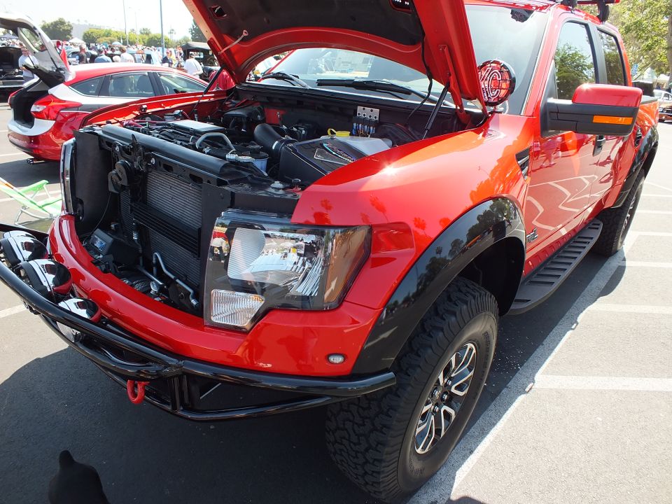 Front three-quarter view of a red and black Ford Raptor with hood open at a car show