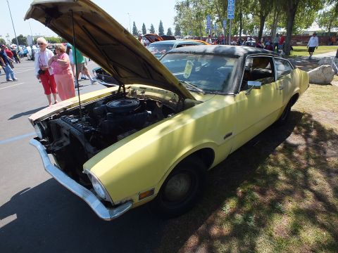 2003 Ford Crown Victoria - faded yellow 1970s Ford Maverick two-door coupe sits with its hood raised, exposing the engine, while spectators stand nearby.